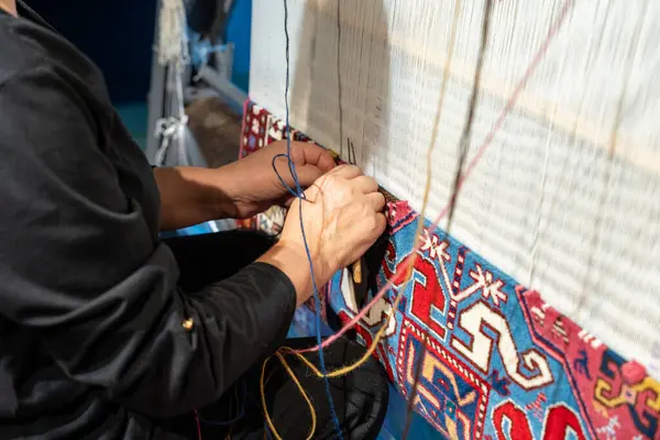 Bir adam halıyı dikmek için iğne iplik kullanıyor. Halı renkli ve çiçek ve yıldızlardan oluşan bir deseni var. Adam işine odaklanmış.