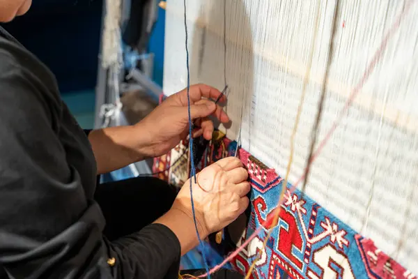 Bir kadın halıda çalışıyor, elleri ipliklerle meşgul. Halı renkli ve çiçek desenli. Kadın işine odaklanmış durumda, ve sahne kendini işine adadığını gösteriyor.