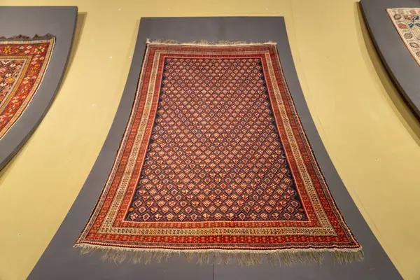 Bir müzede sarı arka planda bir halı sergilenir. Halı, kare şeklinde mavi ve kırmızıdır.