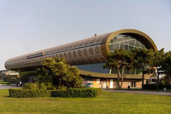 Bir sürü penceresi ve altın yapraklı tasarımı olan büyük bir bina. Bina ağaçlar ve otlarla çevrili.