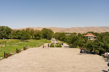Bir grup insan parkta bir taş geçitte yürüyorlar. Gökyüzü açık ve güneş ışıl ışıl parlıyor. Atmosfer huzurlu ve rahatlatıcı, insanlar açık havayı seviyor.