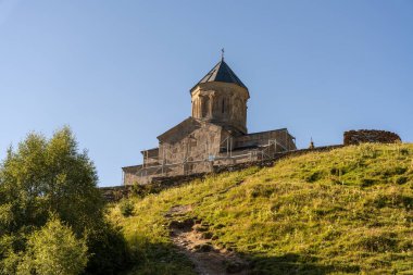 Çan kulesi olan büyük bir taş bina bir tepenin üzerinde duruyor. Bina ağaçlar ve otlarla çevrili. Gökyüzü açık ve mavidir, ve güneş parlıyor. Barış ve sükunet kavramı