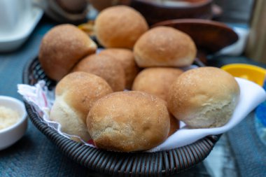 Masanın üzerinde bir sepet ekmek oturuyor. Ekmek kahverengidir ve hafif çıtır bir dokusu vardır. Sepet örüldü ve üzerine kırmızı ve beyaz bir bez örüldü. Ekmek düzgün bir yığın halinde düzenlenir.