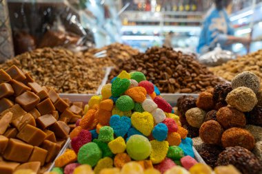 Bir mağazada renkli şeker ve fındık yığını sergileniyor. Şekerler çeşitli şekil ve renklerde ve fındıklar üst üste yığılmış. Sahne canlı ve davetkar.