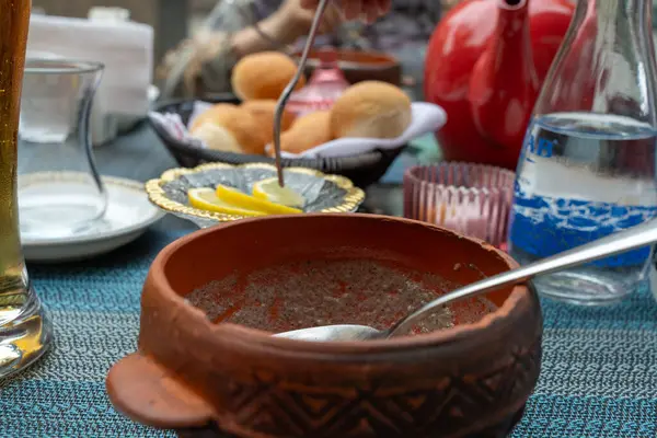 Bir kase yemek içinde kaşık olan bir masada. Kase kahverengi ve üzerinde bir tasarım var. Masada başka kase ve kaşıklar da var.