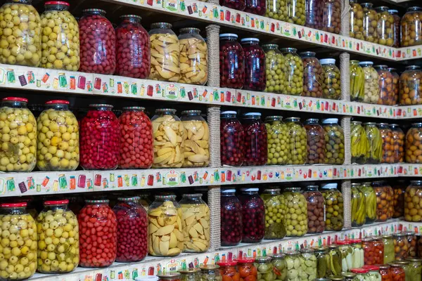 Turşu ve zeytin de dahil olmak üzere çeşitli sebzelerle dolu bir kavanoz. Kavanozlar bir rafta dizilmiş, bazıları diğerlerinden daha uzun. Sahne bolluk hissi veriyor.