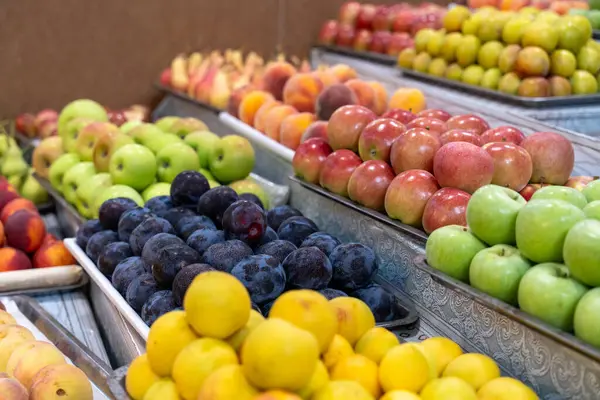 Elma, portakal ve erik gibi çeşitli meyvelerle dolu bir meyve tezgahı. Elmalar farklı renk ve boyutlarda ve portakallar bir köşeye yığılmış. Erikler de sergileniyor.