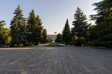 Ortasında çeşme olan büyük boş bir avlu. Avluyu çevreleyen ağaçlar. Gökyüzü mavi ve güneş parlıyor.