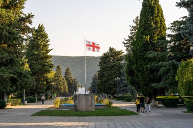 Bir fıskiyenin önünde bir bayrak dalgalanıyor. İnsanlar fıskiyenin ve bayrağın etrafında yürüyor.