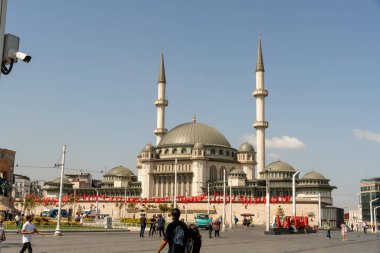 Kırmızı bukleli büyük beyaz bir bina ve mavi gökyüzünün önünde bir kubbe bulunur. Bina kırmızı ve beyaz çizgili bir çitle çevrilidir. Bölgede dolaşan bir sürü insan var.