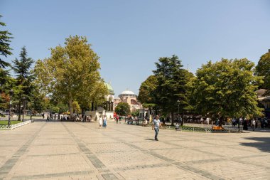 Yürüyen ve banklarda oturan insanlarla dolu bir şehir meydanı. Atmosfer canlı ve hareketli. Arka planda büyük bir binanın varlığı, sahnenin kentsel hissini artırır.