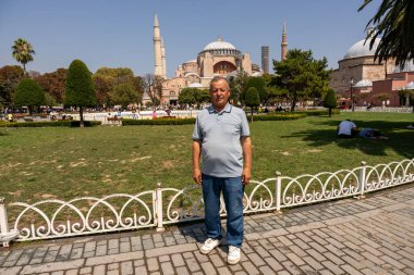 Bir adam büyük bir binanın önünde duruyor. Adam mavi gömlek ve kot pantolon giyiyor.