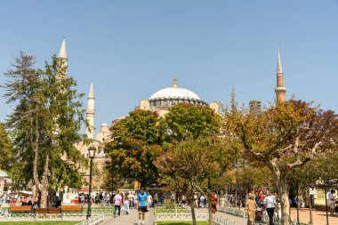 Büyük bir binanın yakınındaki bir parkta büyük bir grup insan yürüyor. Park ağaçlar ve banklarla dolu ve gökyüzü açık ve mavi.