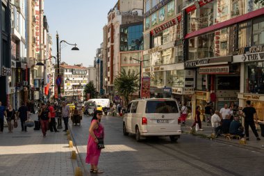 Şehir caddesinde pembe elbiseli bir kadın kaldırımda dikiliyor. Etrafta dolaşan bir sürü insan var ve caddede bir araba sürüyor.