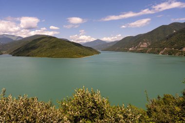 Arkasında dağlar olan güzel bir göl. Su sakin ve berrak. Gökyüzü mavi ve bulutlu.