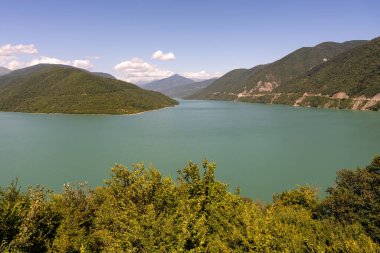 Arkasında dağlar olan güzel bir göl. Su sakin ve berrak. Gökyüzü mavi ve bazı bulutlar var. Huzurlu ve huzurlu bir sahne.