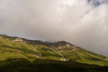 Dağ yeşil çimlerle kaplı ve etrafında sisli bir sis var. Gökyüzü bulutlu ve güneş bulutların arasında parlıyor.