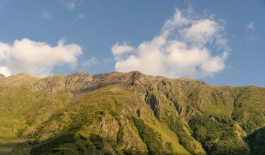 Dağ yeşil çimenler ve ağaçlarla kaplıdır. Gökyüzü açık ve mavidir, birkaç bulut etrafa saçılmıştır. Dağ sırası güzel ve huzurlu bir manzaradır.