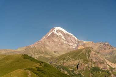 Karla kaplı zirvesi ve açık mavi gökyüzü olan bir dağ. Dağ yeşil çimenler ve ağaçlarla çevrilidir.