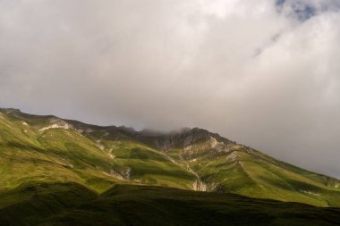 Dağ yeşil çimlerle kaplı ve etrafında sisli bir sis var. Gökyüzü bulutlu ve güneş bulutların arasında parlıyor.