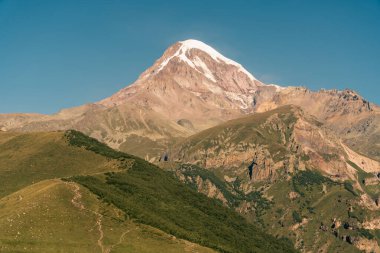 Karla kaplı zirvesi ve altında yeşil bir vadisi olan bir dağ. Gökyüzü açık ve mavi