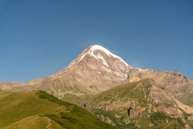 Karla kaplı zirvesi ve açık mavi gökyüzü olan bir dağ. Dağ yeşil çimenler ve ağaçlarla çevrilidir.