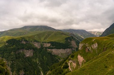 Dağlar yemyeşil ağaçlarla kaplı ve gökyüzü bulutlu. Huzurlu ve huzurlu bir manzara. Dağlar ihtişam ve ihtişam hissi veriyor. Bulutlar derinlik katıyor
