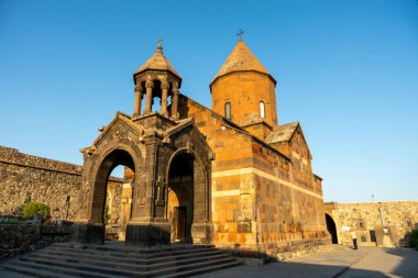 Çan kulesi ve tepesinde haç olan büyük bir bina. Bina taştan yapılmış ve bir sürü kemeri var. Gökyüzü mavi ve güneş parlıyor.