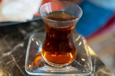 Masadaki kare bir tabakta bir bardak çay duruyor. Çay kahverengidir ve bardak berraktır.
