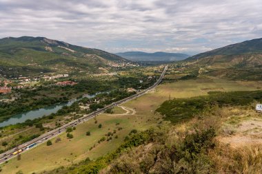 İçinden otoyol geçen bir dağ sırası. Otoyol araba ve kamyonlarla dolu. Gökyüzü bulutlu ve manzara çoğunlukla düz.