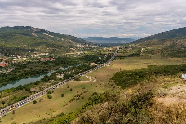 İçinden otoyol geçen bir dağ sırası. Otoyol araba ve kamyonlarla dolu. Gökyüzü bulutlu ve manzara çoğunlukla düz.