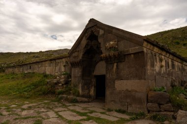Küçük bir girişi olan büyük bir taş bina. Bina eski ve taş bir çatısı var. Gökyüzü bulutlu ve çimenler yeşil