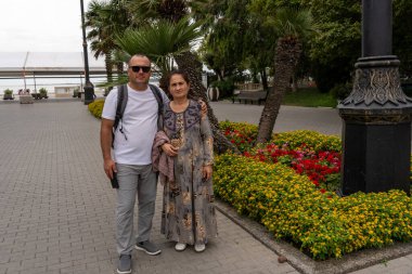 Bir adam ve bir kadın elinde çiçeklerle bir ağacın önünde duruyorlar. Kadın elbise giyiyor ve adam beyaz gömlek giyiyor.