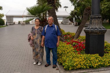 Bir adam ve bir kadın elinde çiçeklerle bir ağacın önünde duruyor. Adam mavi gömlek ve kot pantolon giyiyor.