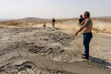 Bir adam yerdeki büyük bir deliğin önünde duruyor. Adam çıplak göğüslü ve kot pantolon giyiyor. Delik kayalık bir alanla çevrili.