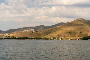 Önünde göl olan güzel bir dağ sırası. Göl sakin ve huzurlu, üzerinde birkaç tekne yüzüyor. Dağlar ağaçlarla kaplı ve gökyüzü bulutlu. Sahne huzur dolu.