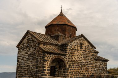 Çan kulesi ve üzerinde haç olan küçük bir kilise. Bina taştan yapılmıştır ve kırsal bir görünüme sahiptir. Gökyüzü bulutlu ve güneş görünmüyor.