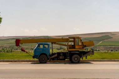 Yolun kenarına mavi ve sarı bir vinç park edilmiş. Vinç eski ve paslıdır ve bir üzüm tarlası ile çevrilidir. Sahne sessiz ve huzurlu.