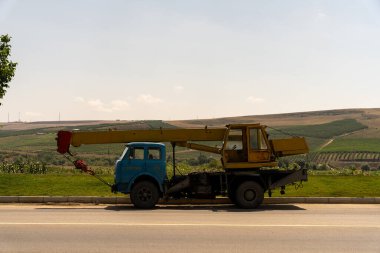 Yolun kenarına park edilmiş sarı ve mavi bir kamyon var. Kamyon bir vinç ve bir tarlaya park edilmiş.