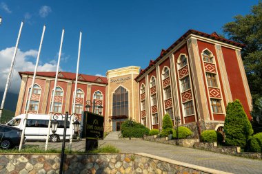 Önünde beyaz bir minibüs olan büyük kırmızı bir bina. Binanın bir sürü penceresi ve geniş bir girişi var.