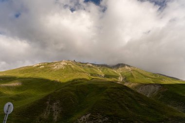 Arka planda bulutlu bir gökyüzü olan bir dağ sırası. Bulutlar beyaz ve kabarık ve dağ yeşil çimlerle kaplı. Huzurlu ve huzurlu bir sahne.