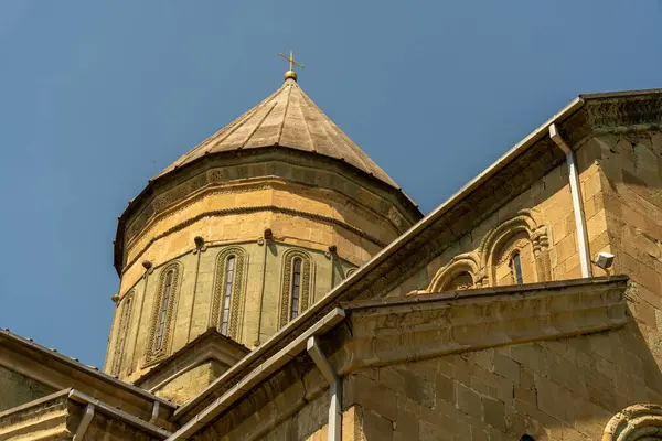 Binanın tepesinde bir çan kulesi ve haç var. Bina kahverengi ve bir sürü penceresi var.