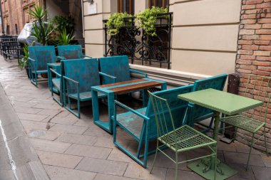 Bir sıra mavi sandalye ve masalar binanın dışında. Sandalyeler sıraya dizildi, bazıları binaya bakıyor, diğerleri de sokağa bakıyor.