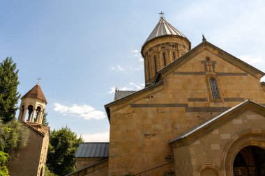 Çan kulesi ve üzerinde haç olan büyük kahverengi bir bina. Bina ağaçlarla çevrili.