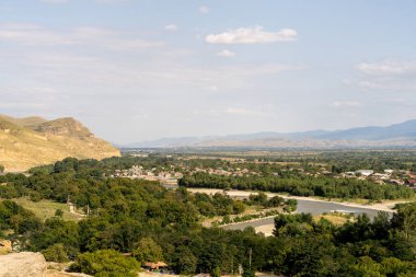 İçinden nehir akan bir dağ sırası. Nehir ağaçlar ve evlerle çevrilidir. Gökyüzü açık ve güneş parlıyor.
