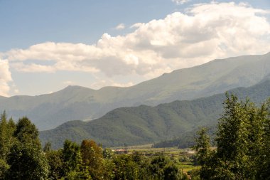 Dağlar ağaçlarla kaplı ve gökyüzü bulutlu. Dağlar çok yüksek ve ağaçlar yeşil.