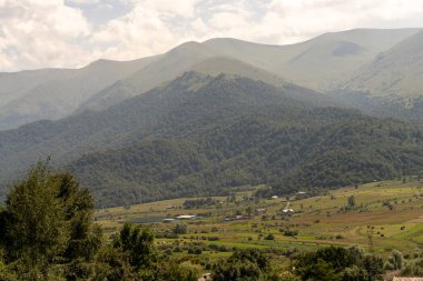 Yeşil yemyeşil ormanı ve vadide küçük bir köyü olan bir dağ sırası. Gökyüzü bulutlu ve dağlar ağaçlarla kaplı. Sahne huzurlu ve sakin, dağlarla birlikte.