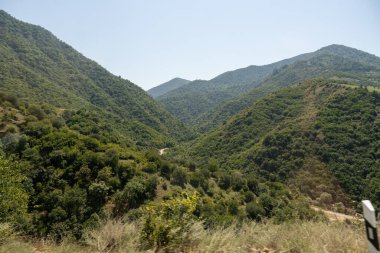 Dağlar yemyeşil ağaçlarla kaplıdır ve gökyüzü açık ve mavidir. Sahne huzurlu ve sakin, dağlar güzel bir zemin sağlıyor.