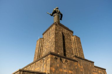 Kılıç tutan bir adamın heykeli yüksek bir binanın üzerindedir. Gökyüzü açık ve mavi ve heykel tuğla bir duvarla çevrili.
