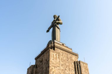 Bir binanın tepesinde gitar tutan bir kadın heykeli var. Gökyüzü açık ve mavi, ve heykel taş bir duvarla çevrili.